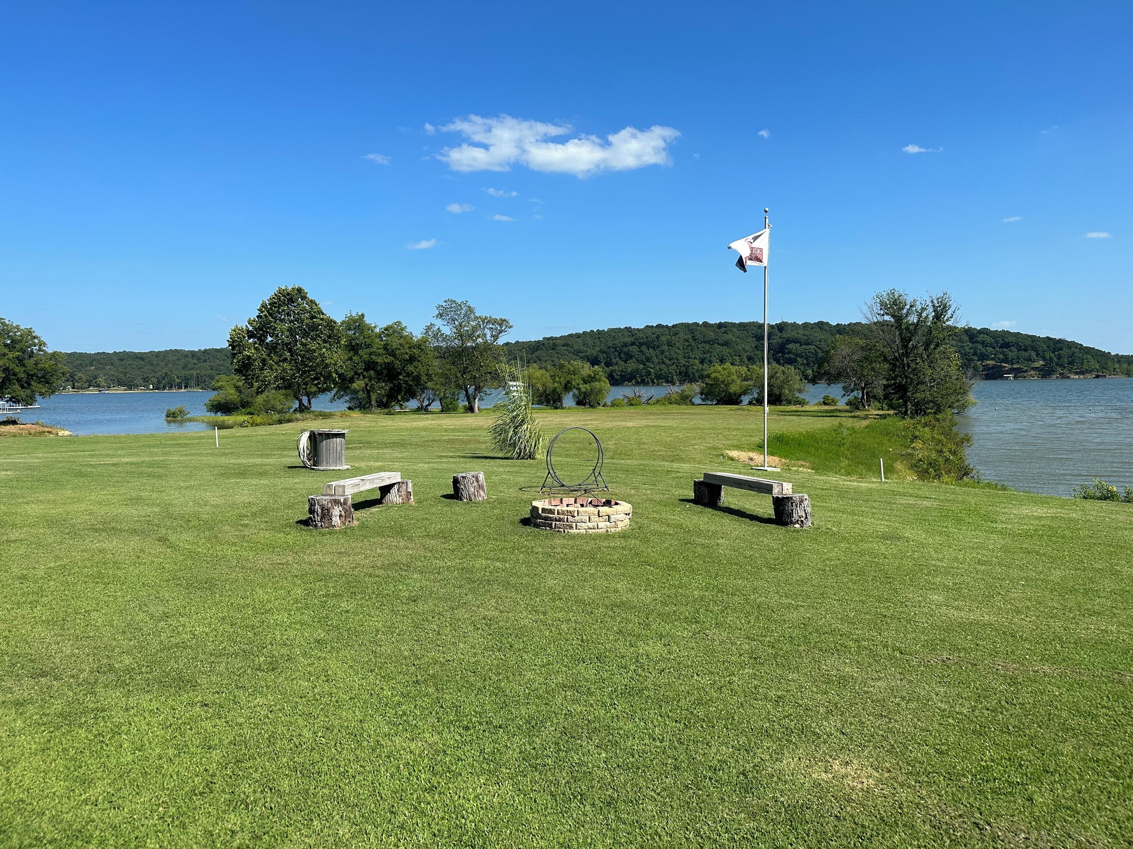 A grassy area with benches and a flagpole, surrounded by trees and a lake.