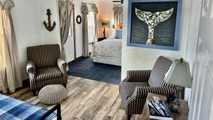 Une chambre d’hôtel avec des fauteuils à rayures, un pouf capitonné, une œuvre d’art représentant la queue d’une baleine et un lit avec une tête de lit à motifs floraux.