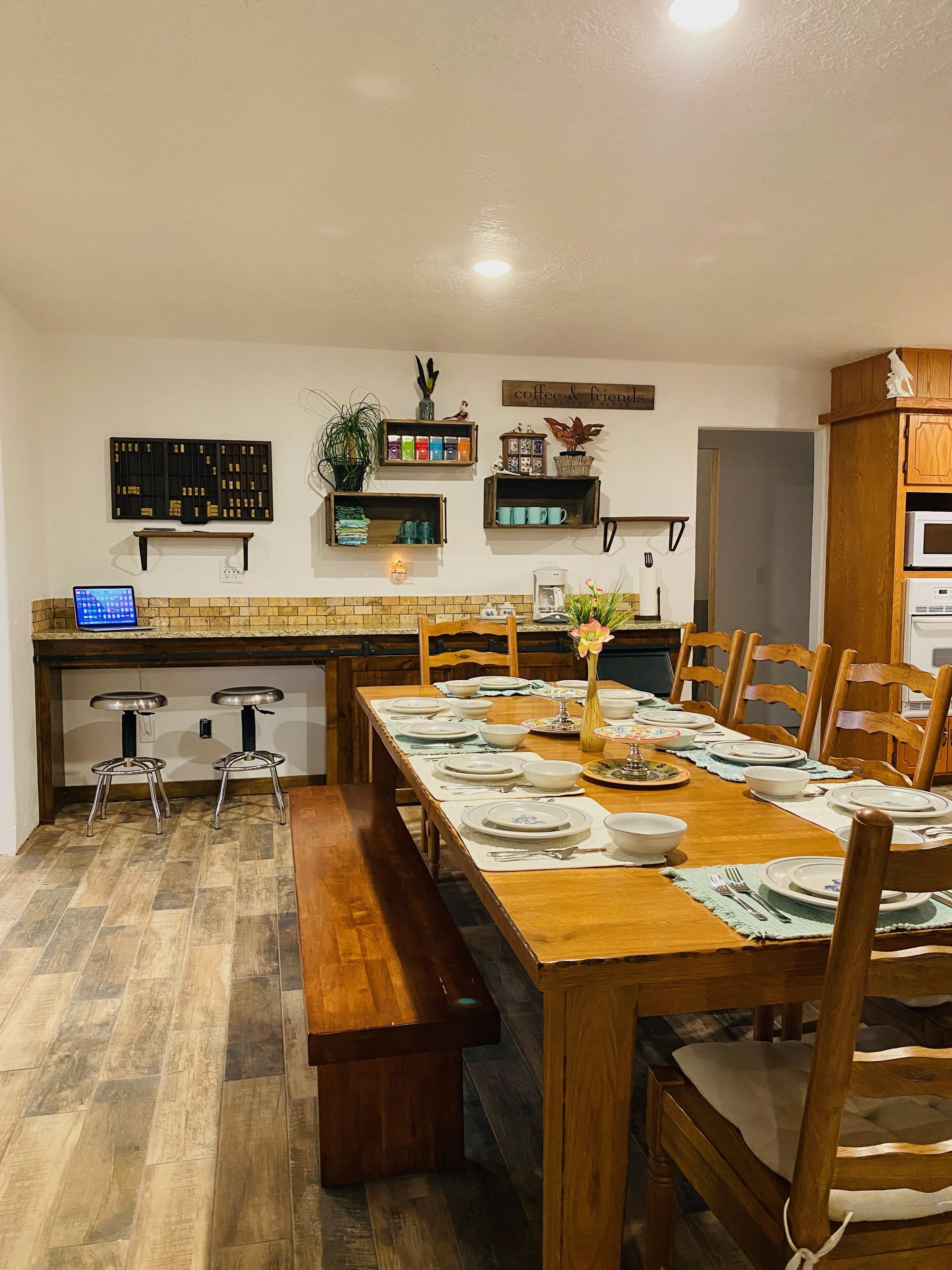 Un coin repas avec une table en bois dressée pour six personnes, entourée de chaises en bois. Un banc est placé le long d’un des côtés de la table. La pièce comprend un coin cuisine avec un comptoir et un four à micro-ondes. On peut voir des étagères remplies de divers objets et une pancarte sur laquelle est écrit « café et amis » sur le mur.