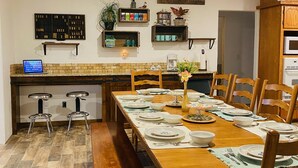 Un coin repas avec une table en bois dressée pour six personnes, entourée de chaises en bois. Un banc est placé le long d’un des côtés de la table. La pièce comprend un coin cuisine avec un comptoir et un four à micro-ondes. On peut voir des étagères remplies de divers objets et une pancarte sur laquelle est écrit « café et amis » sur le mur.