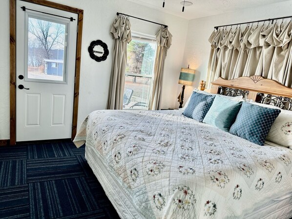 Un dormitorio con una cama grande, cabecera de madera, espejo, puerta y ventana con cortinas.