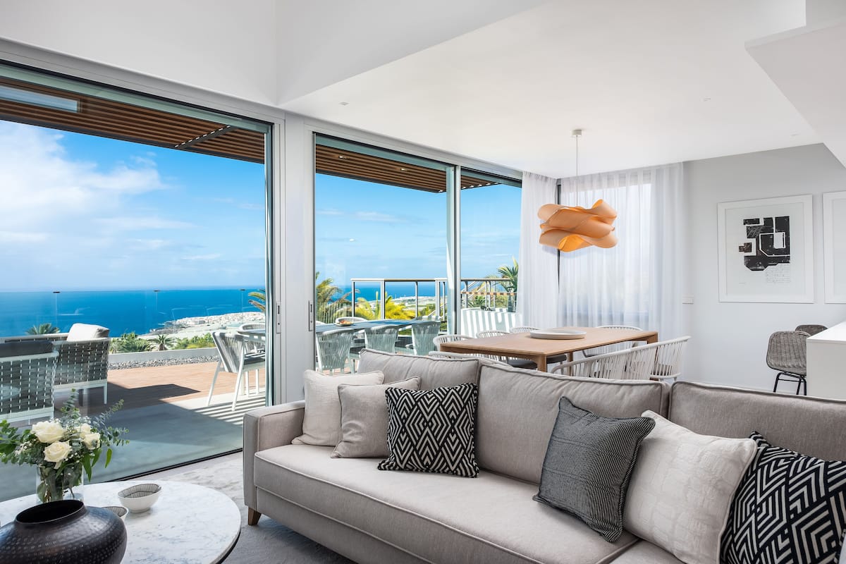 A modern living room with a large sofa, a dining table, and a view of the ocean.