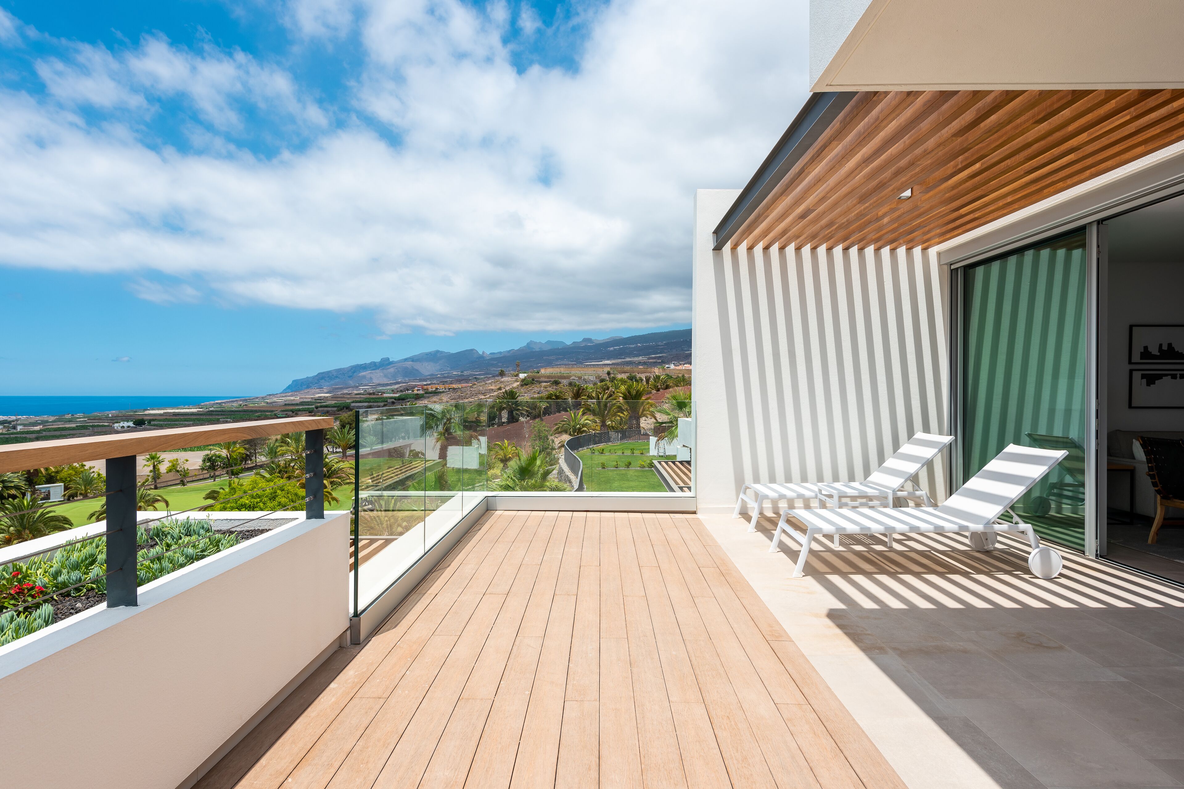 A balcony with a wooden floor, white lounge chairs, and a glass railing overlooking a landscape with greenery and distant mountains.