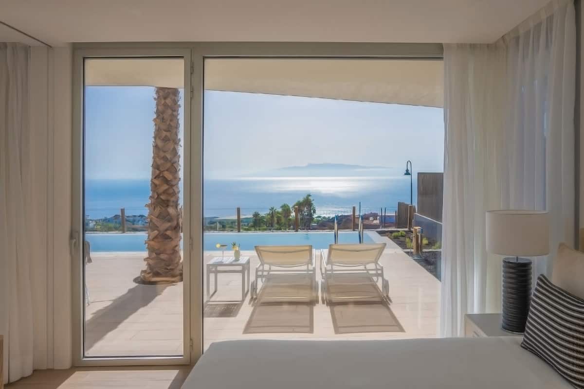A hotel room with a large sliding glass door leading to a balcony with a pool and a view of the ocean.