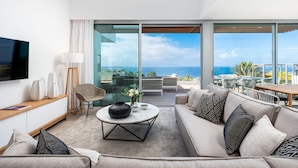Una sala de estar moderna con sofá, mesa de centro y televisor. Cuenta con ventanas grandes con vista al océano.