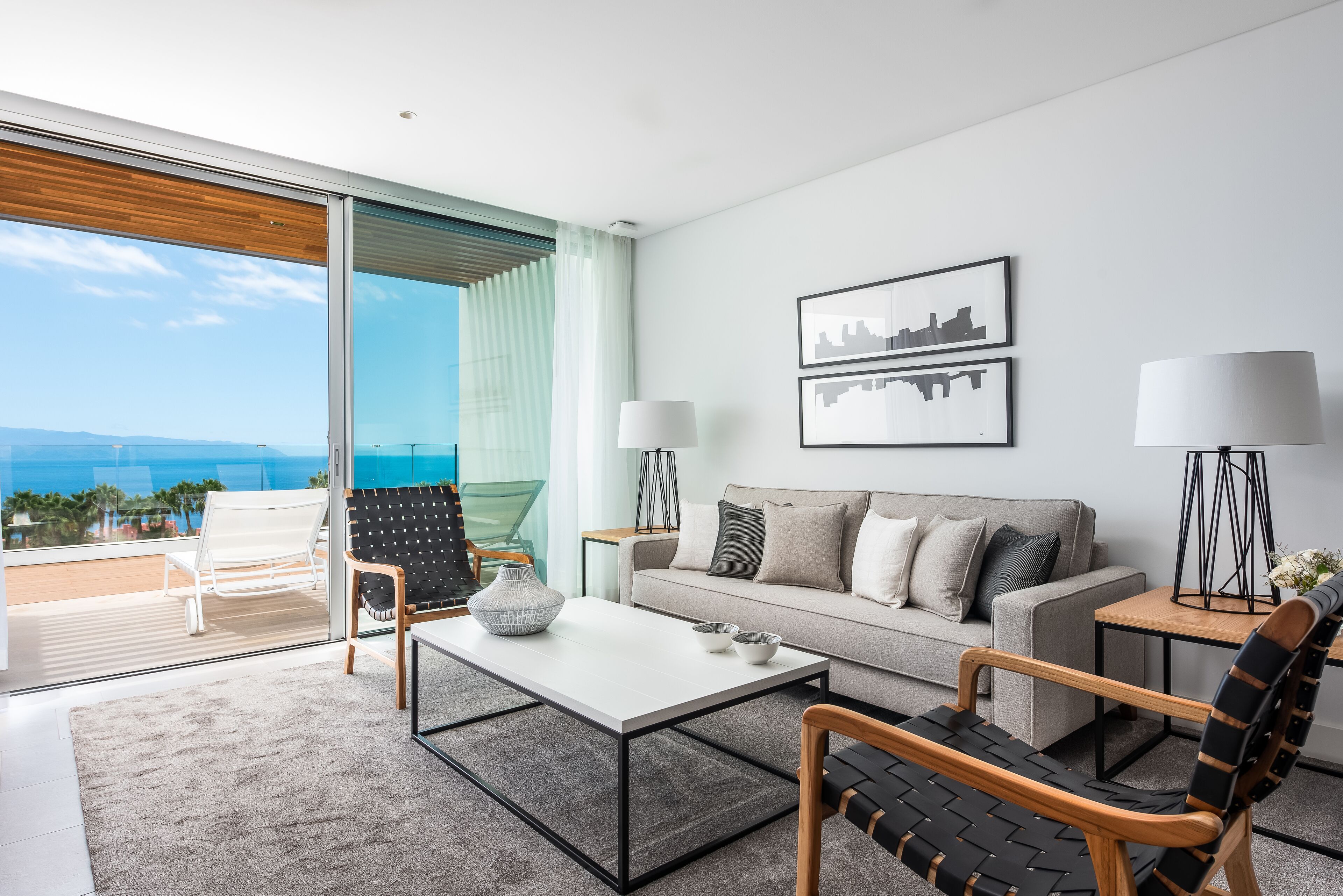 Un salón moderno con sofá, mesa de centro y sillones, un ventanal con vistas al océano, y dos lámparas de pared.