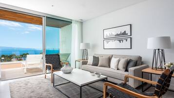 Un salón moderno con sofá, mesa de centro y sillones, un ventanal con vistas al océano, y dos lámparas de pared.