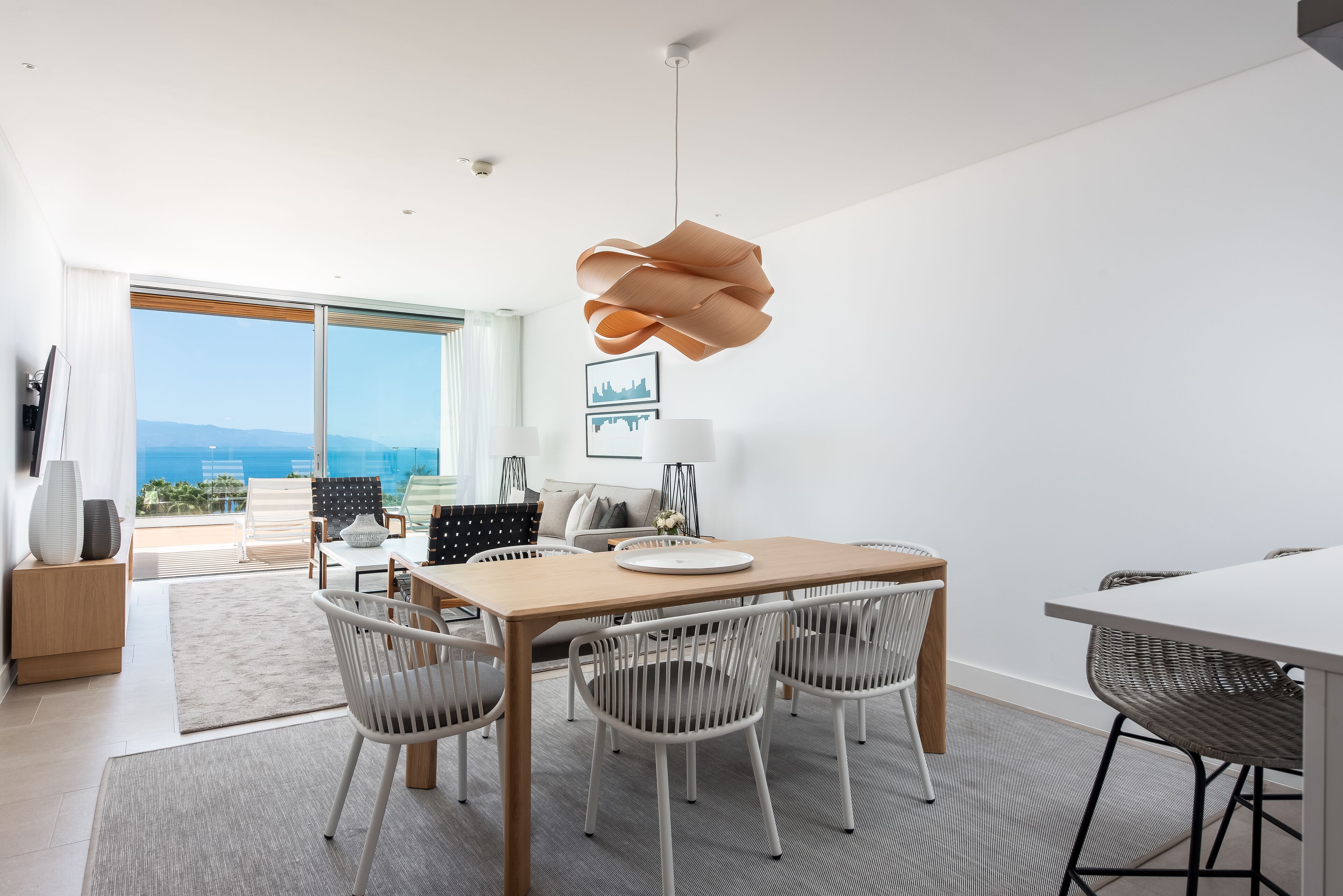 Un comedor moderno con una mesa de madera y sillas blancas, una lámpara colgante original y vistas al océano a través de amplios ventanales.
