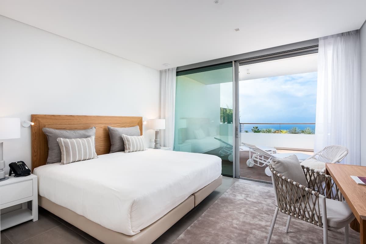 A hotel room with a large bed, a bedside phone, a chair, and a table.