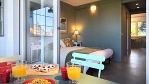 Ein Hotelzimmer mit Bett, Esstisch mit Frühstücksangebot, Fenster mit Blick auf die Stadt und einem Gang zu einem weiteren Zimmer.