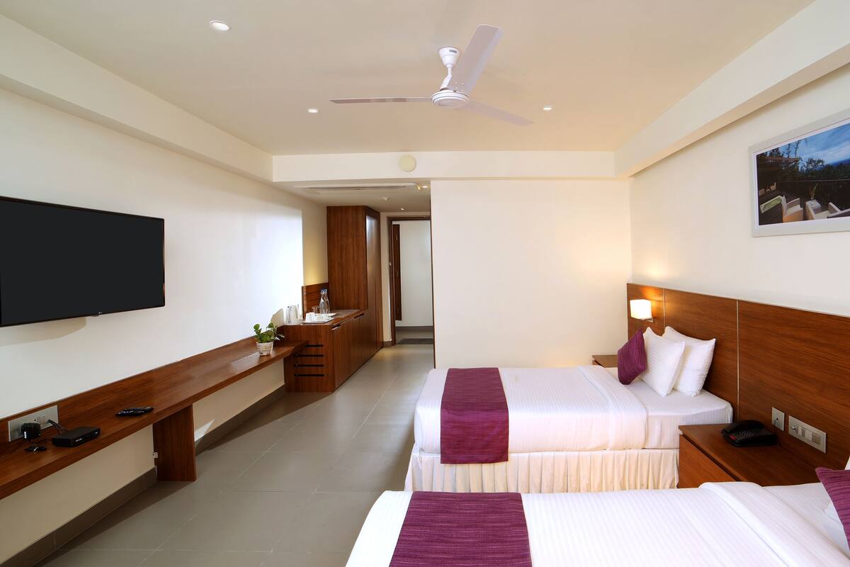 Habitación de hotel con dos camas, televisor, escritorio y ventilador de techo.