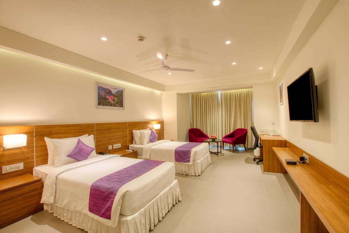A hotel room with two beds, a desk, a TV, a ceiling fan, and a window with curtains.