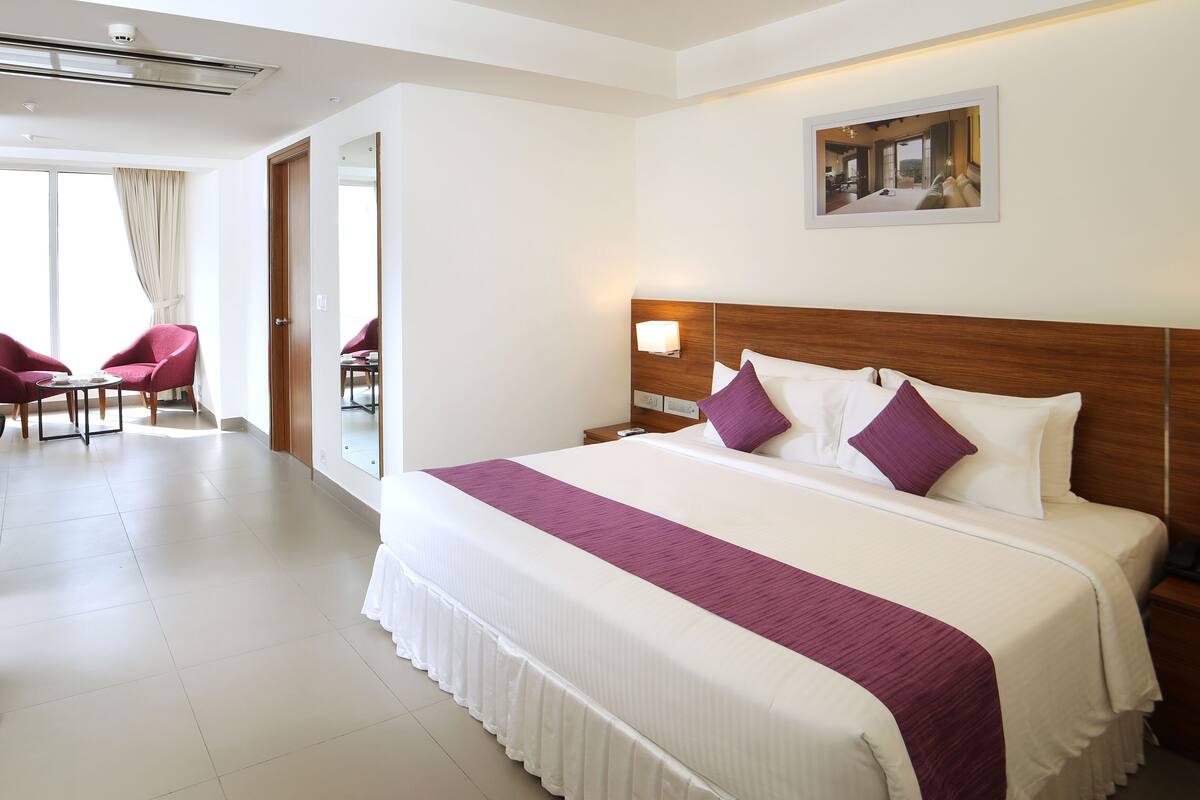 Habitación de hotel con una cama grande, cabecera de madera, mesita de noche, silla, mesa y una ventana amplia.