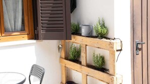 A wooden outdoor seating area with a table and chairs, a window with brown shutters, and a small shelf with plants and a mug. - The Pearl of Neptune Tourist rental 600mt from the sea (Nettuno)