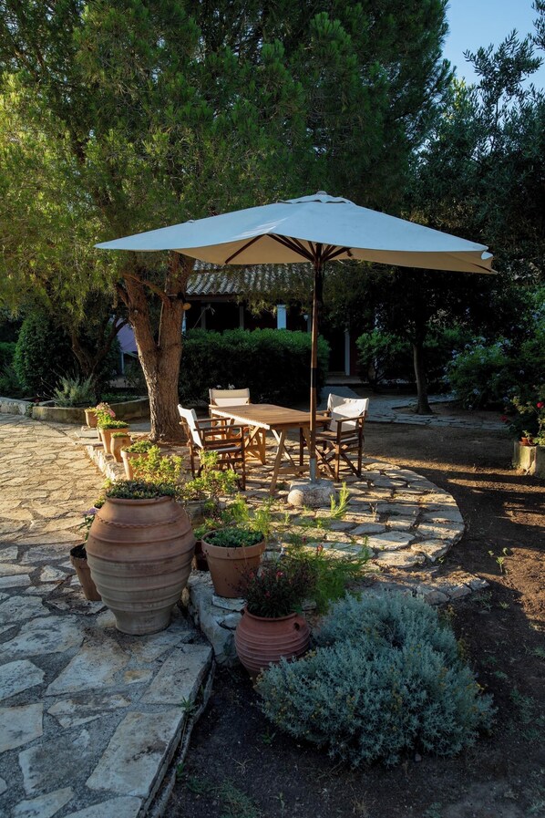 Un patio al aire libre con una mesa y sillas de madera bajo un amplio paraguas, rodeado de plantas en macetas y un camino de piedra.