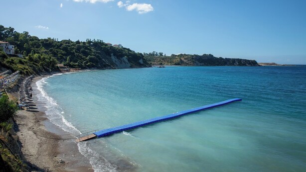 A coastal beach with a long blue boardwalk extending into the sea, surrounded by lush greenery and rocky cliffs.