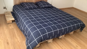 A bedroom with a wooden headboard, a bed with a dark blue checkered bedspread, and wooden pallets under the bed.