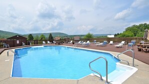 A rectangular swimming pool with a metal ladder on one side, surrounded by lounge chairs and tables on a wooden deck.
