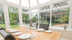 Un invernadero con una mesa de comedor dispuesta para cuatro personas, rodeado de amplios ventanales que ofrecen vistas a un jardín y una casa.