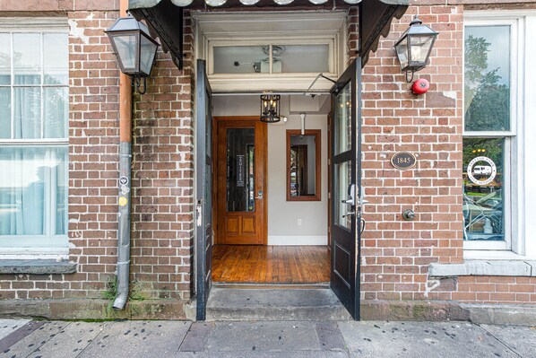 A brick building entrance with a wooden door, a small porch, and a window with a bicycle outside.