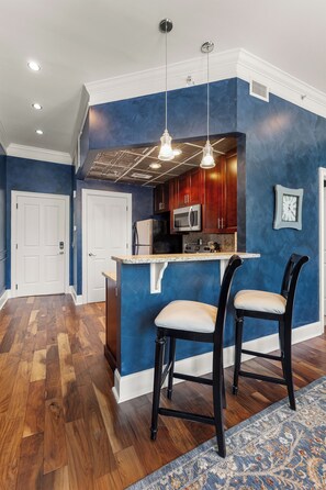 A modern kitchen with a blue accent wall, wooden cabinets, a stainless steel microwave, and two bar stools.