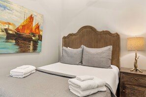A neatly made bed with pillows, a painting of boats on the wall, a lamp, and a stack of towels. - Charming Downtown Studio One Block From The River by Lucky Savannah (Savannah)