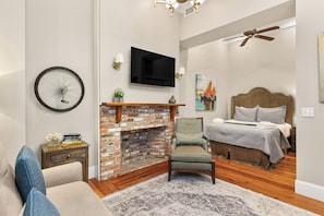 A bedroom with a brick fireplace, a television mounted on the wall, a bed with a headboard, and a ceiling fan.