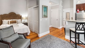 A bedroom with a bed, nightstand, and chair, a hallway leading to a kitchen with a refrigerator and bar stool. - Charming Downtown Studio One Block From The River by Lucky Savannah (Savannah)