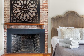 Une chambre avec une cheminée en briques, une tête de lit en bois et un lit avec une literie blanche et des coussins à motifs.
