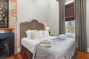 A neatly made bed with pillows and a blanket, a bedside lamp, a window with curtains, and a brick wall.