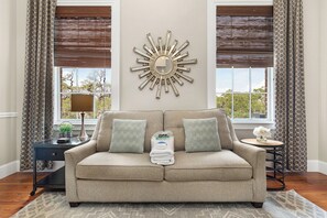 Una sala de estar con un sofá color beige, dos almohadones estampados, un espejo decorativo y vistas al exterior.