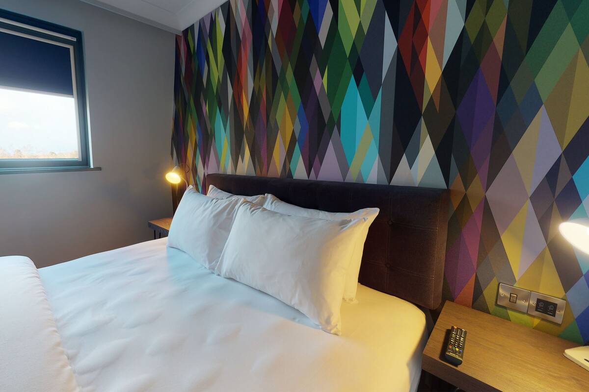A hotel room with a bed, pillows, a nightstand, a remote control, and a wall with a geometric pattern.