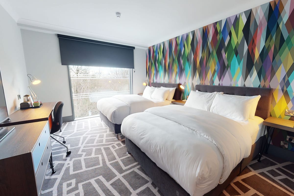 A hotel room with a large bed, a desk, and a chair. There is a window with a view of trees.