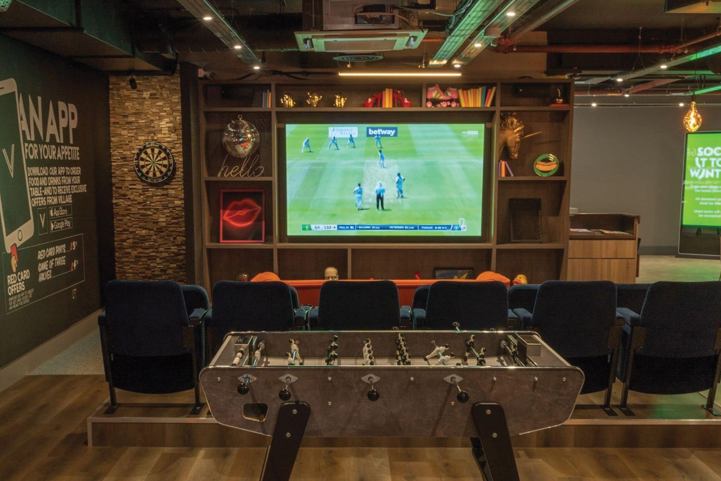 Una habitación con mesa de futbolín, dardos y un televisor mostrando un partido de fútbol.
