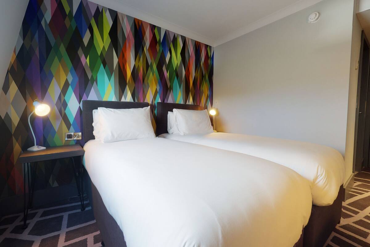 A hotel room with a bed, two bedside tables, and a colorful geometric-patterned wall.