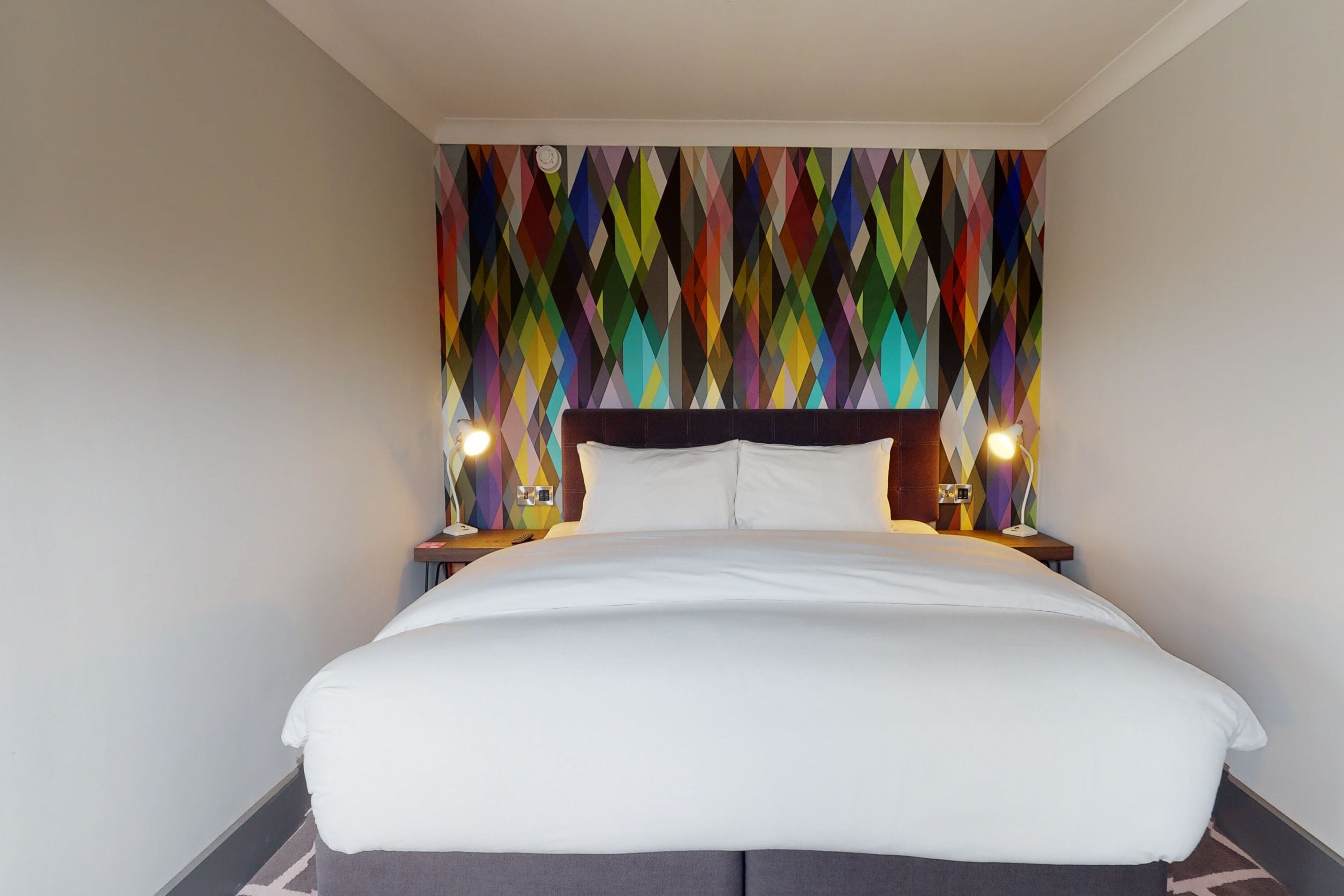 A hotel room with a large bed, a colorful geometric wall design, and two bedside lamps.