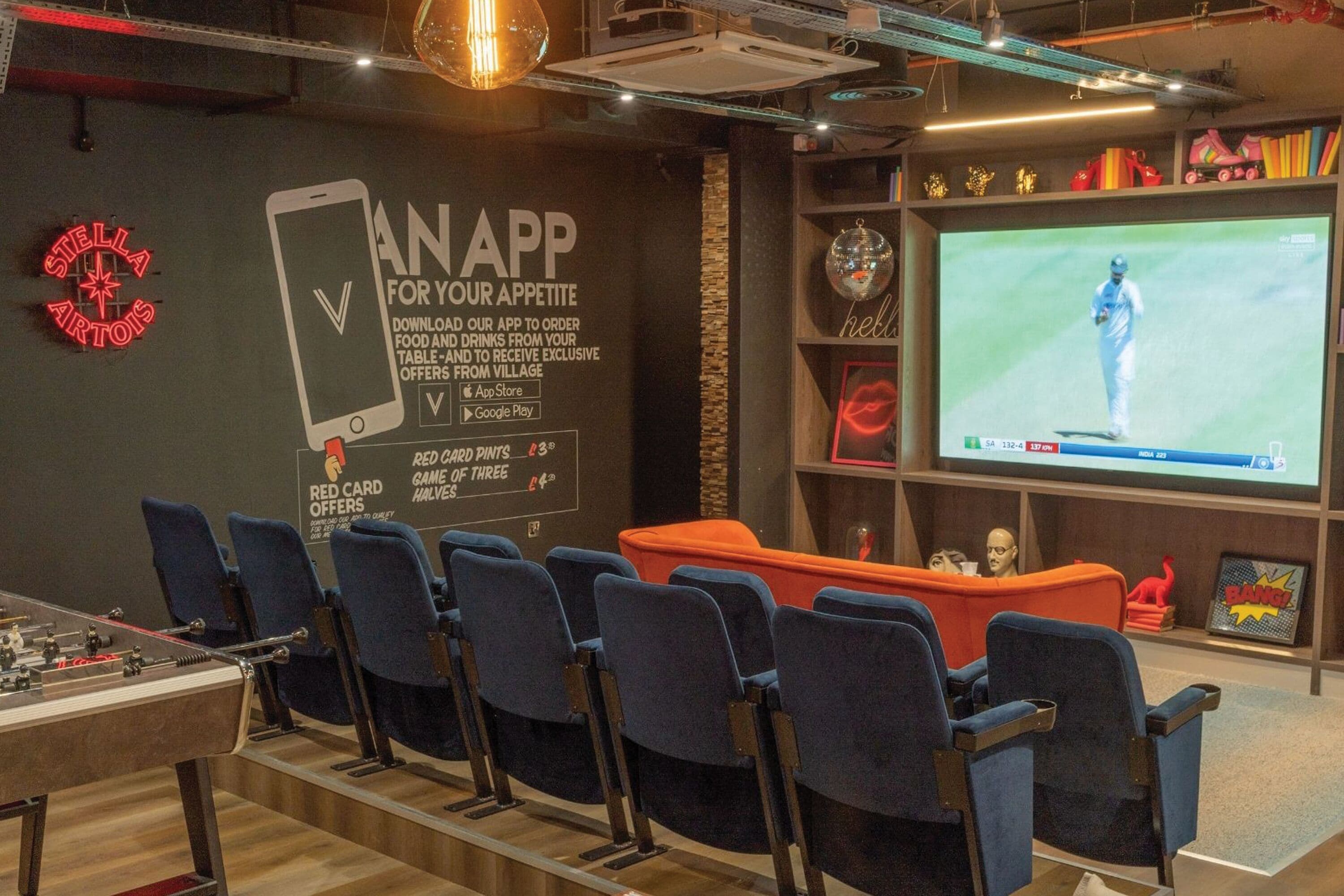 A modern lounge area with a foosball table, comfortable seating, a large screen for viewing sports, and a neon sign.