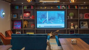 A modern lounge area with blue sofas, a wooden table, and a large TV screen displaying a sports event. - Village Hotel Bracknell (Bracknell)