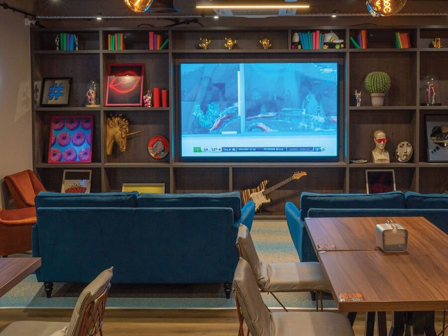 A modern lounge area with blue sofas, a wooden table, and a large TV screen displaying a sports event.