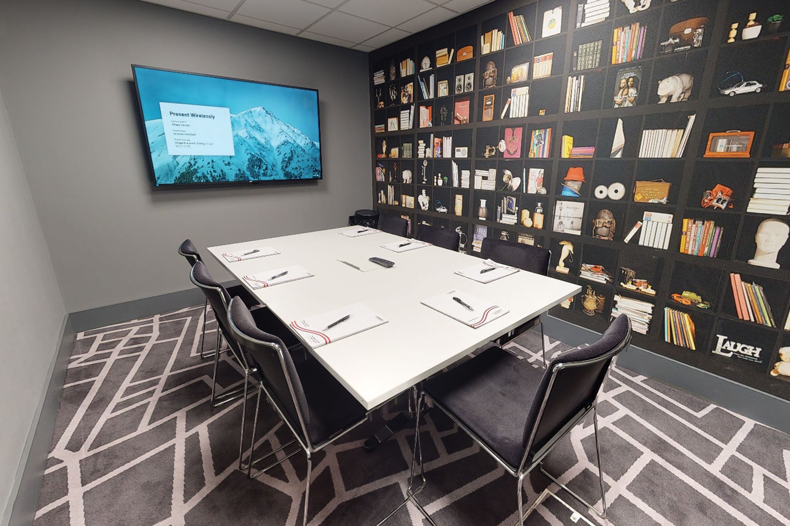 A conference room with a large table, chairs, a screen displaying text, and a bookshelf with various items.