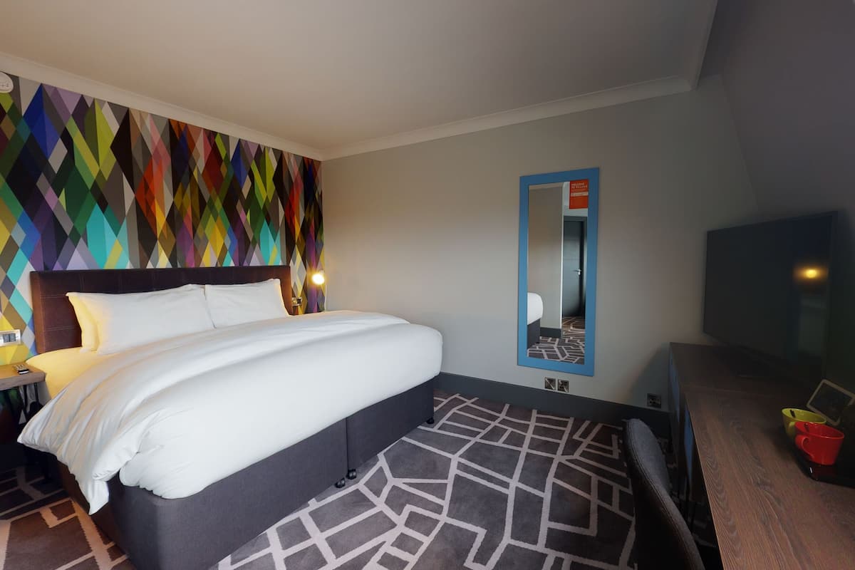 A hotel room with a large bed, a colorful geometric wall design, a mirror, and a TV.