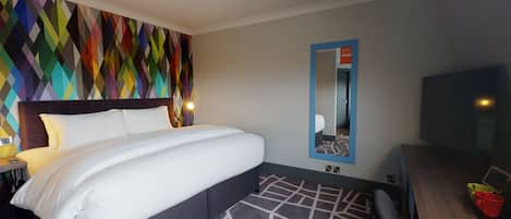 Quarto de hotel com uma cama grande, design geométrico colorido na parede, um espelho e uma televisão.
