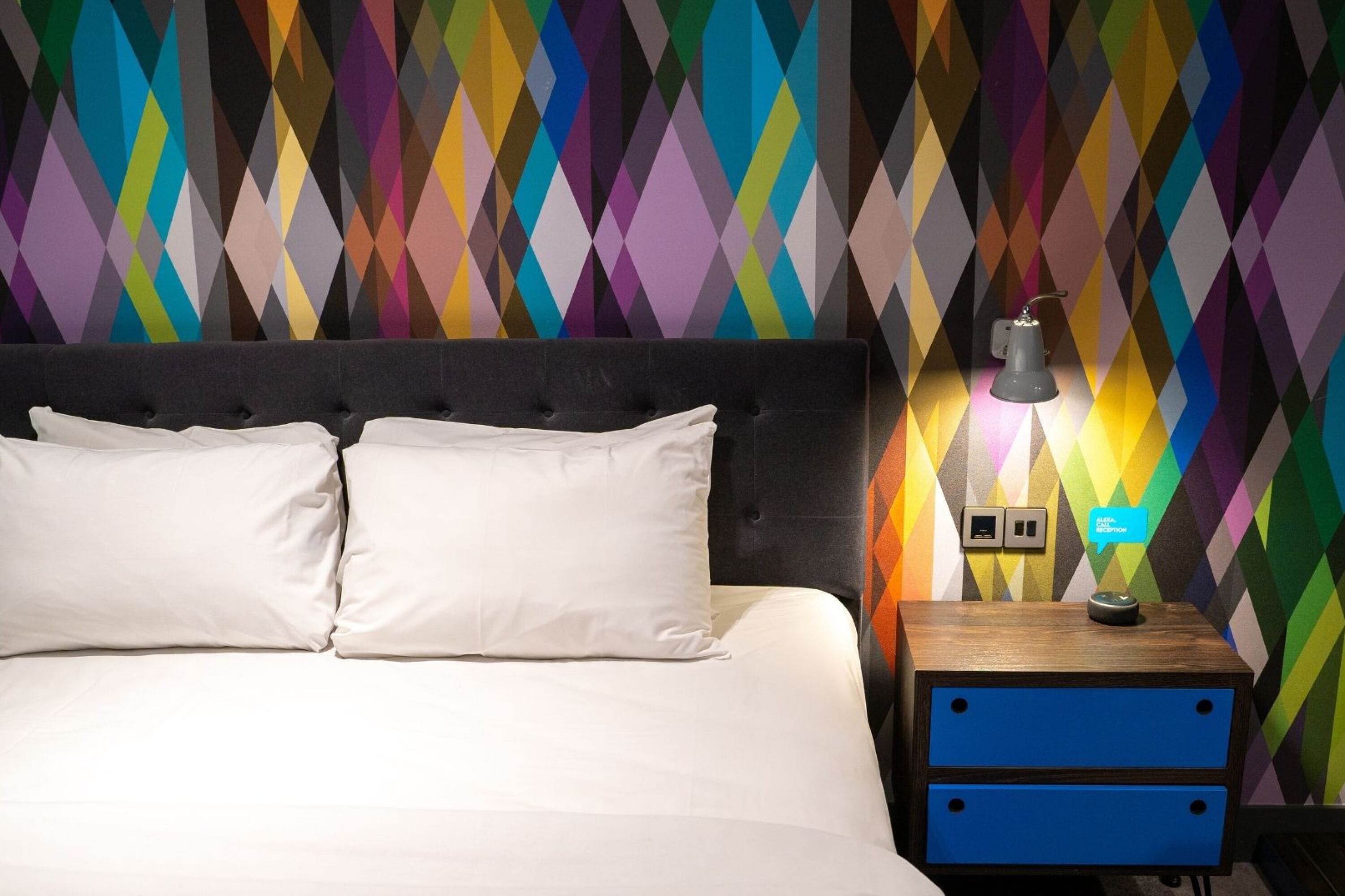 A bed with white linens and two pillows, a nightstand with a lamp, and a colorful diamond-patterned wall.