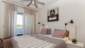 Ein Schlafzimmer mit Bett, Nachttischen, Deckenventilator, einem Fenster mit Vorhängen, einem Wandbild und einer Topfpflanze.