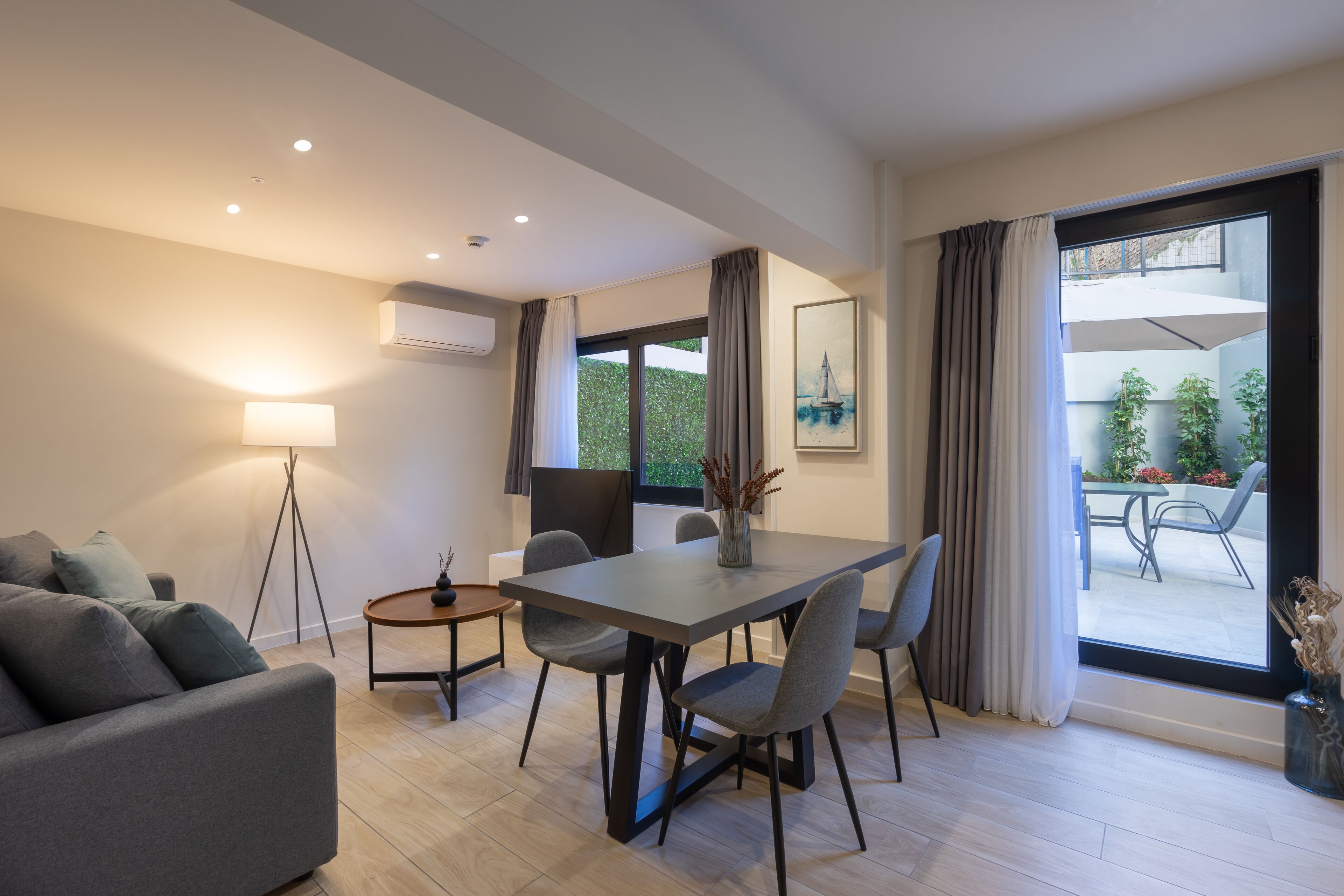 A modern living room with a dining table, grey sofas, a floor lamp, and a view of the outdoors through large windows.