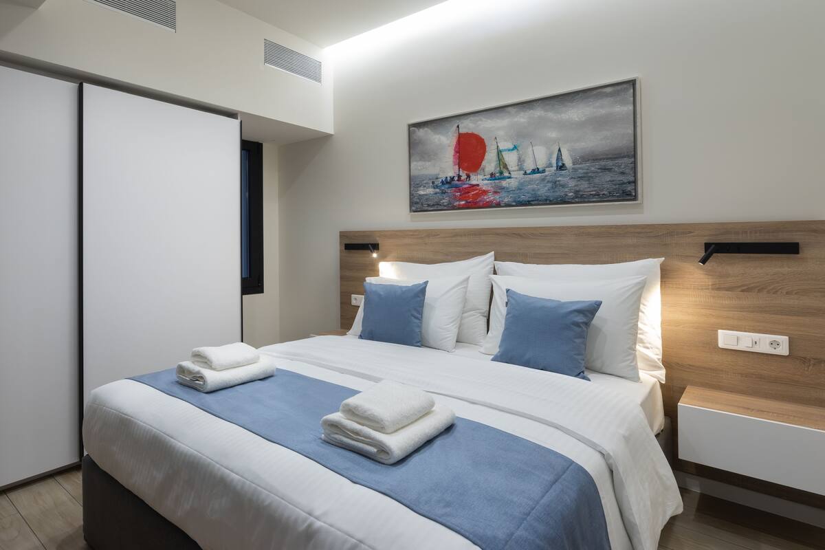 A neatly made bed with blue and white bedding, a painting of sailboats on the wall, and a wooden headboard.