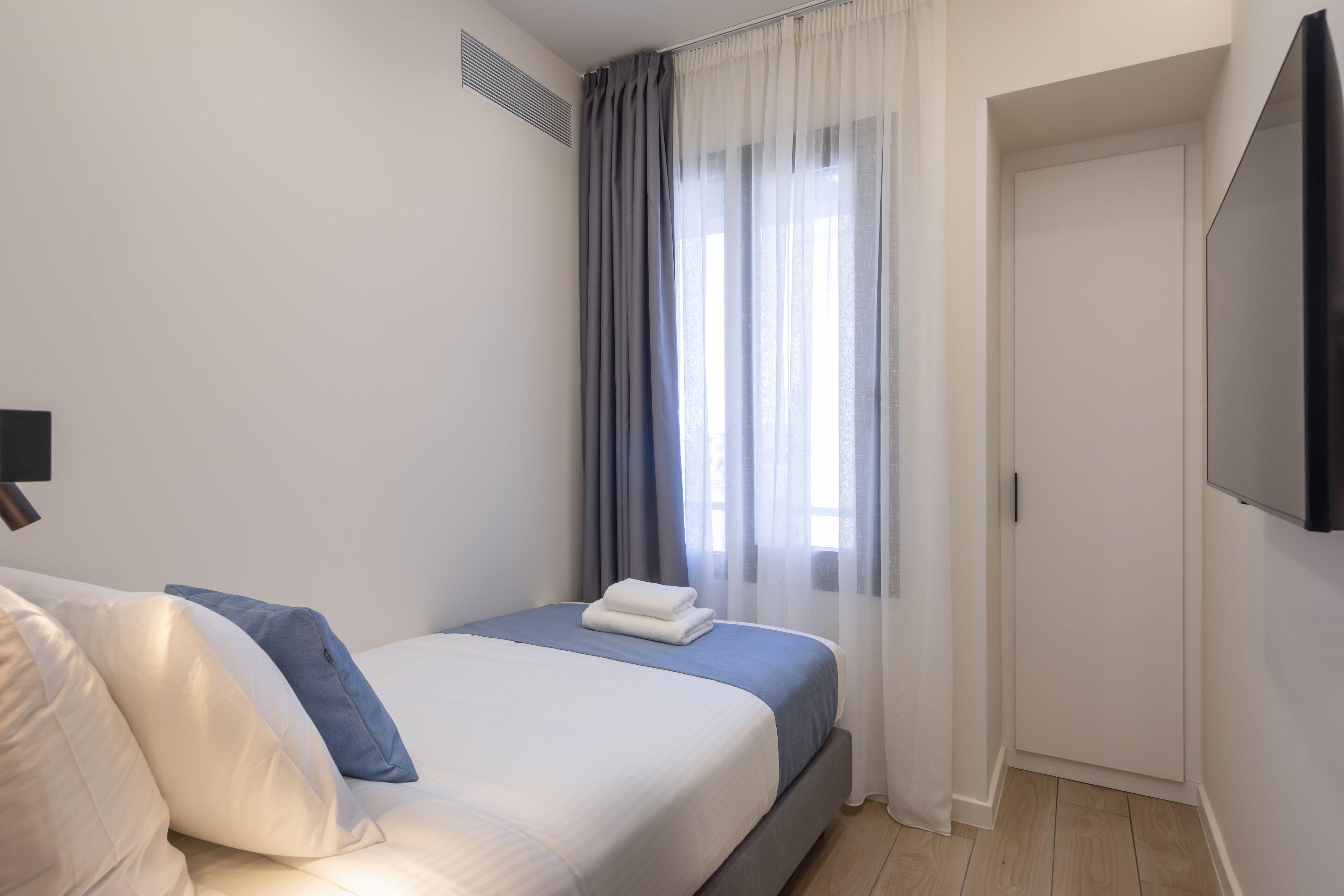 A neatly arranged bedroom with a bed, a flat-screen TV mounted on the wall, and a door leading to another room.