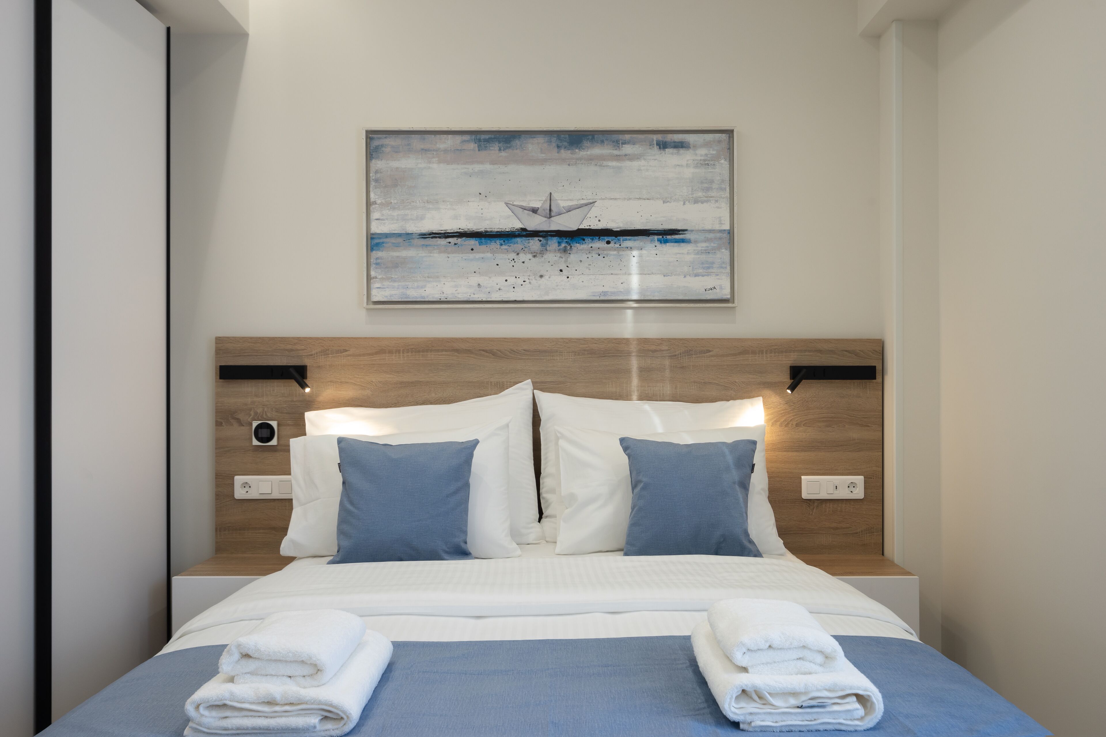 Una cama bien tendida con ropa de cama azul y blanca, un cabecero de madera y un cuadro de un velero en la pared de arriba.
