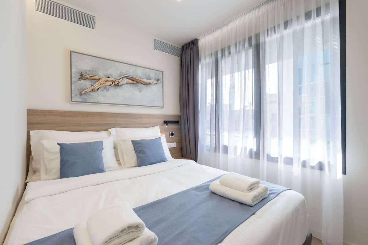 A neatly made bed with white and blue bedding, a framed artwork above it, and a window with sheer curtains.