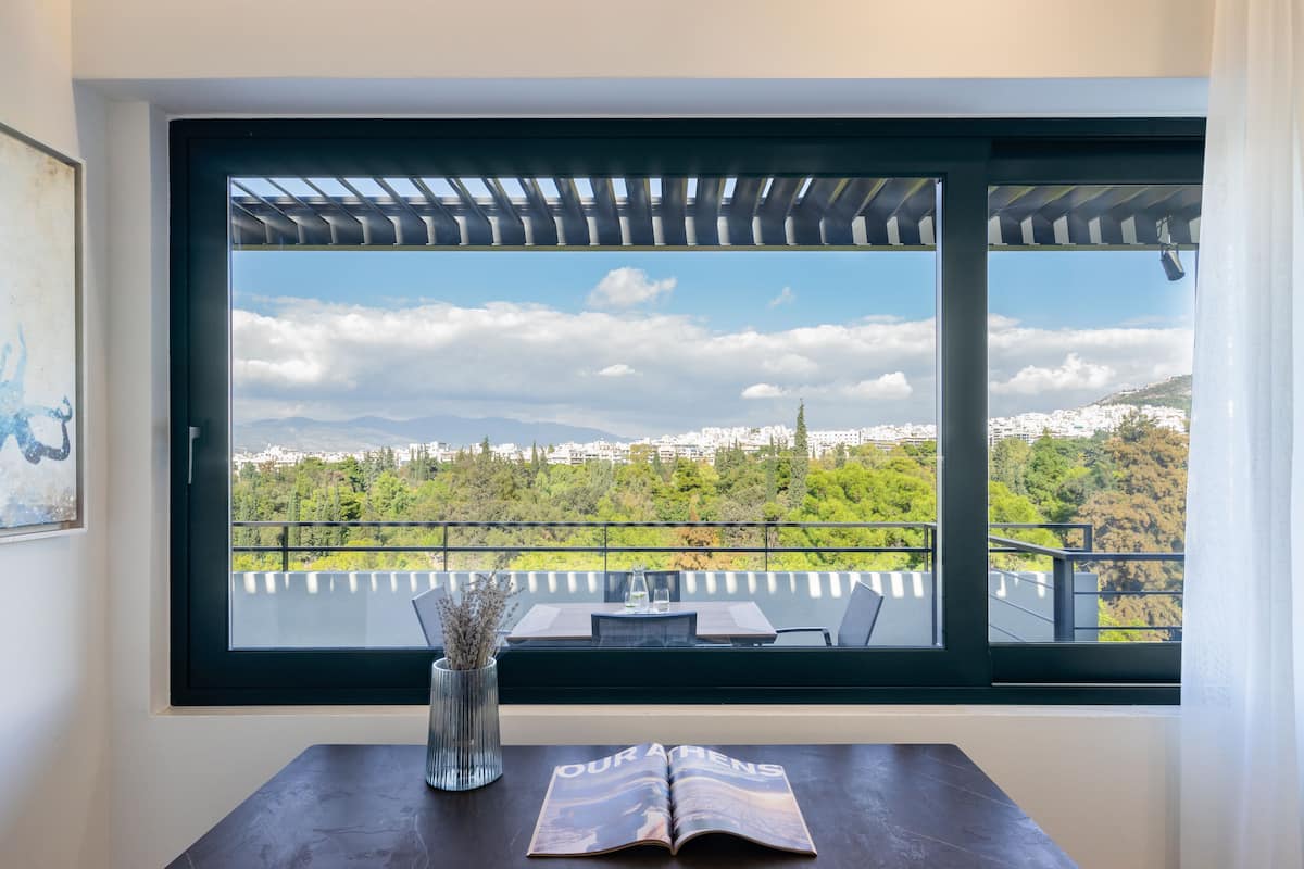 A room with a large window overlooking a scenic view of a city and mountains. There is a table with a vase and an open magazine.